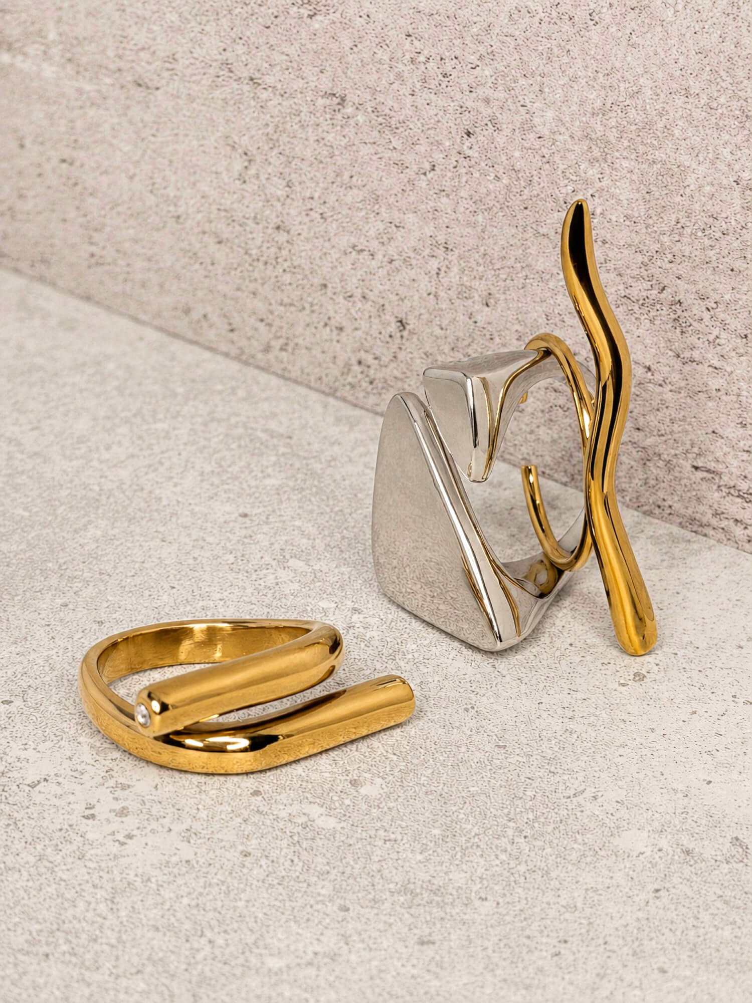 Silver triangle wrap ring, gold wave curve ring, and gold sculptural wrap ring with crystal styled on gray background